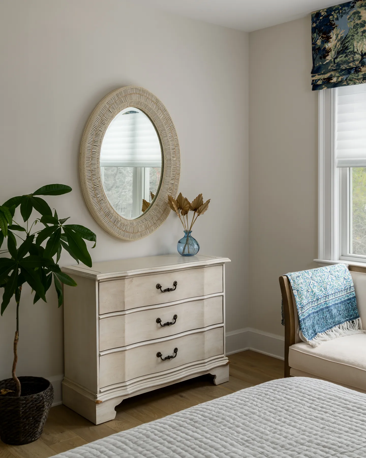 Weathered dresser with oval wicker mirror, blue glass vase, and toile roman shade