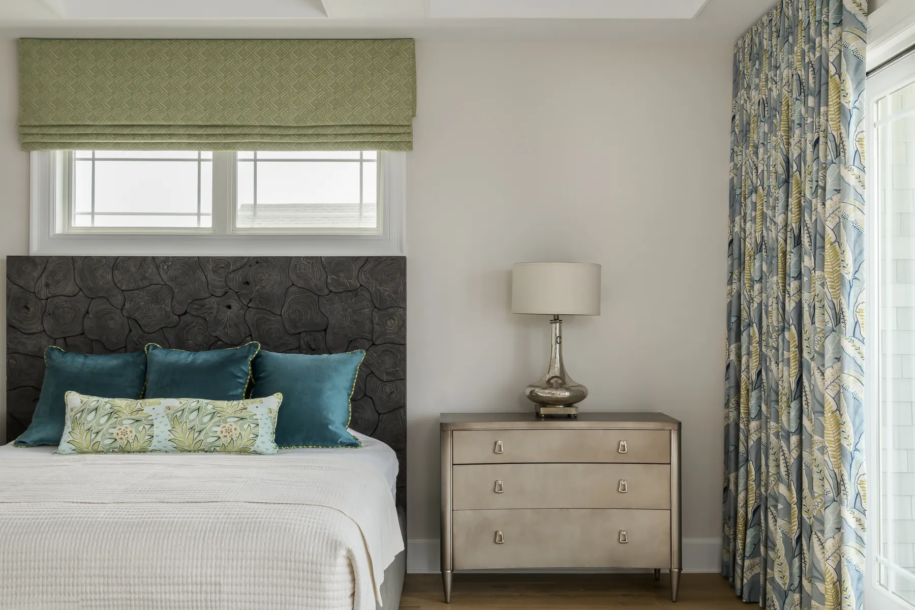 Charcoal headboard detail with teal velvet pillow, glass lamp, and botanical leaf curtain