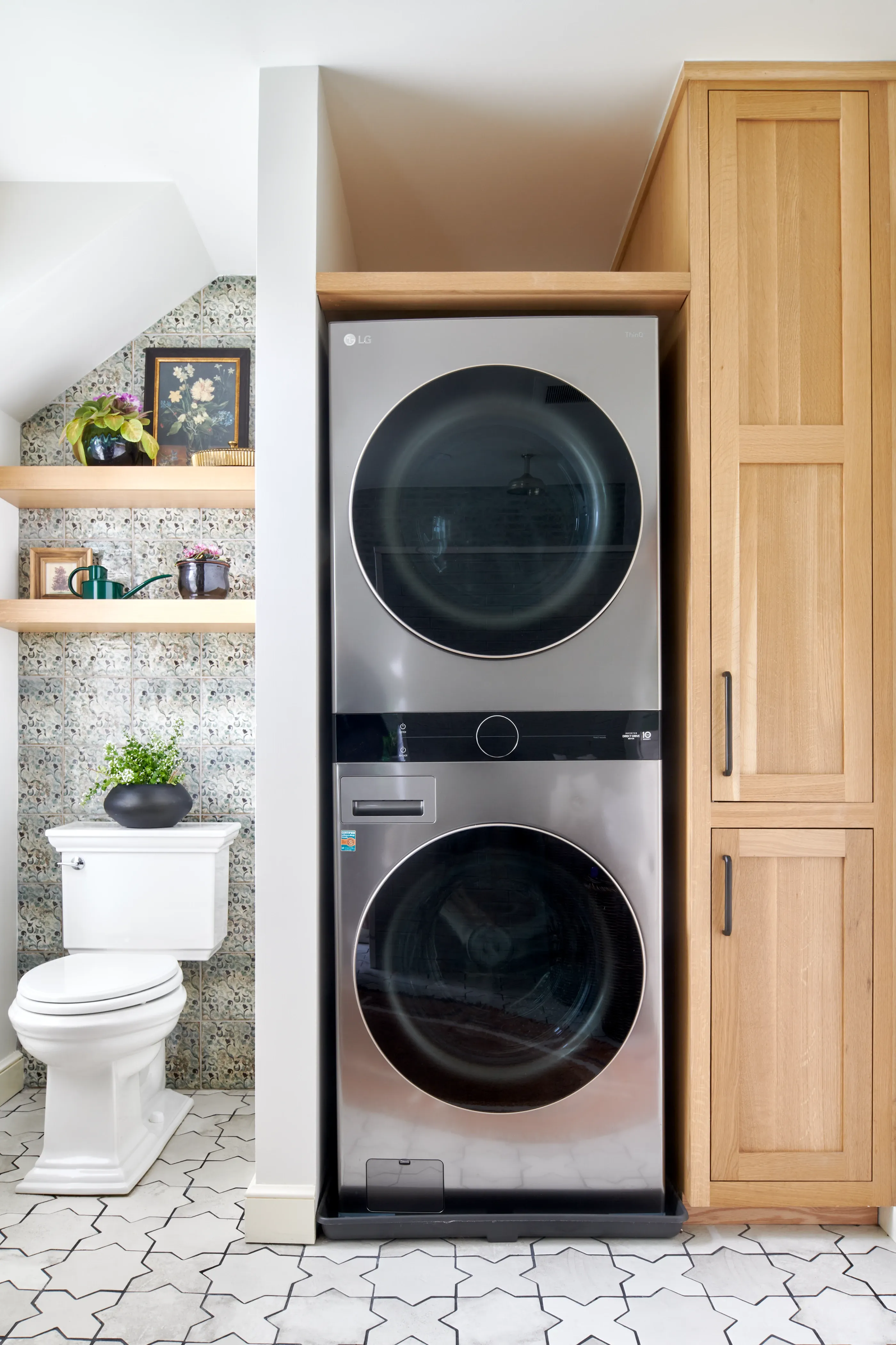 Laundry alcove with stacked units, natural oak cabinetry, and floral wallpaper powder room beyond