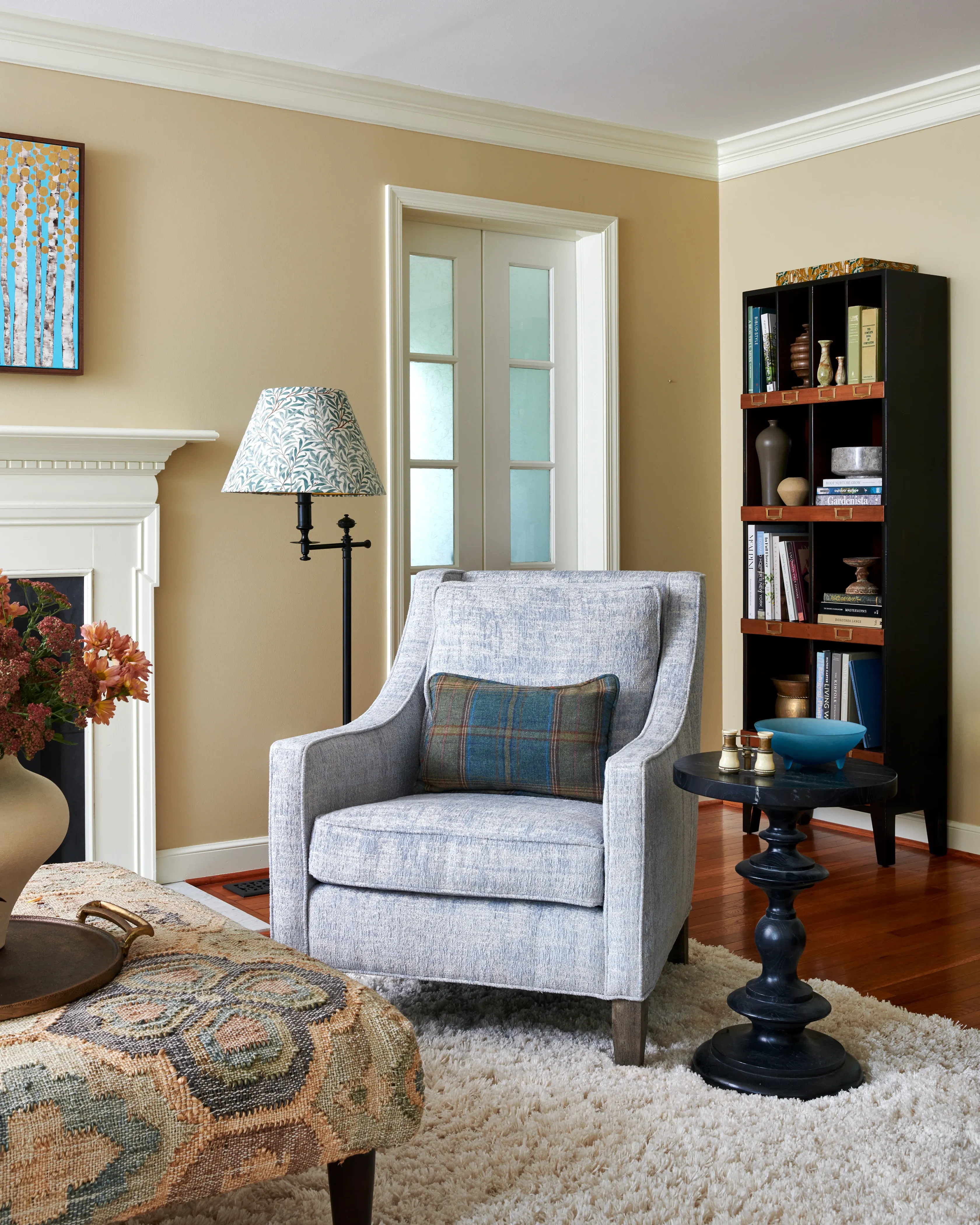 Gray linen club chair with plaid accent pillow, patterned lampshade, and dark bookcase with collected objects